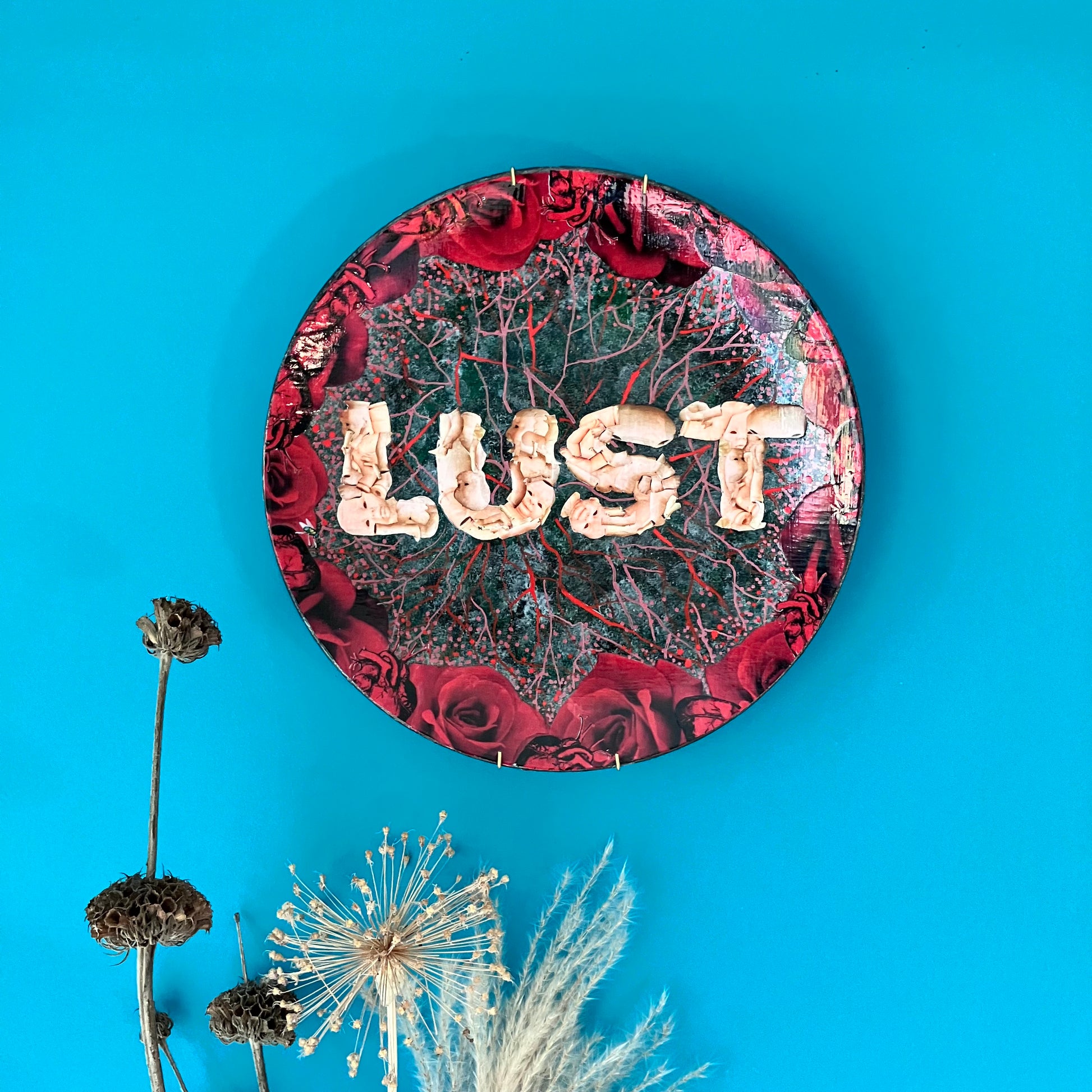 "Lust" Wall Plate by House of Frisson, featuring the word "lust" written with plastic doll parts, framed by red roses and anatomical human heart ,on a charcoal grey background with vein patterns. Lifestyle image of plate on a blue wall.