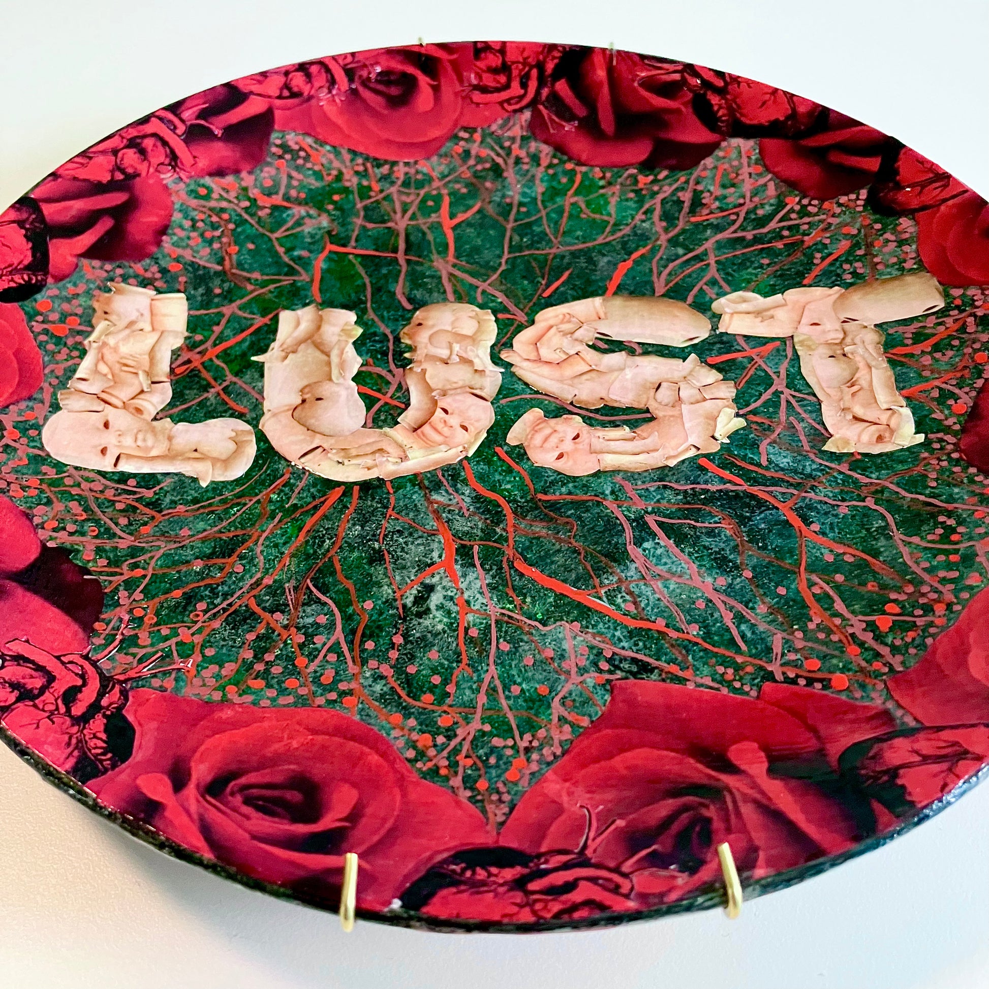 "Lust" Wall Plate by House of Frisson, featuring the word "lust" written with plastic doll parts, framed by red roses and anatomical human heart ,on a charcoal grey background with vein patterns. Closeup details.