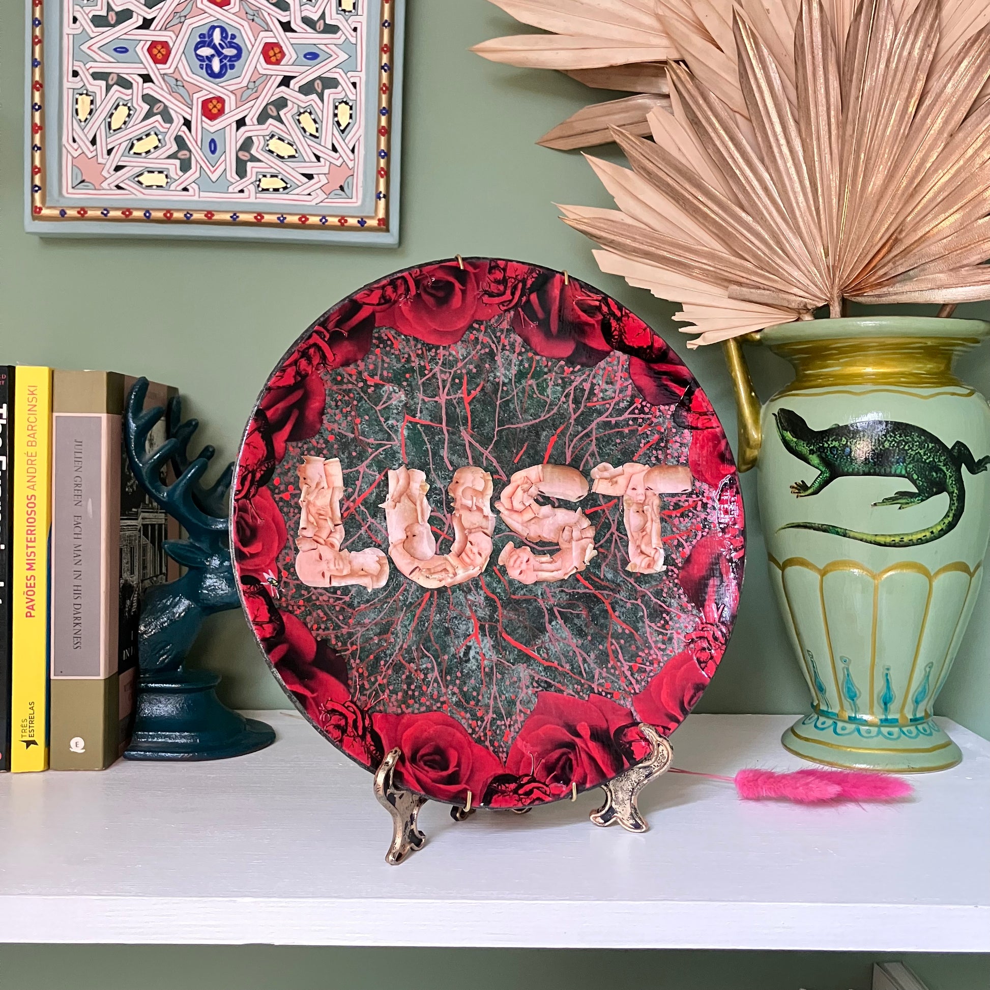 "Lust" Wall Plate by House of Frisson, featuring the word "lust" written with plastic doll parts, framed by red roses and anatomical human heart ,on a charcoal grey background with vein patterns. Plate on a plate stand resting on a shelf.
