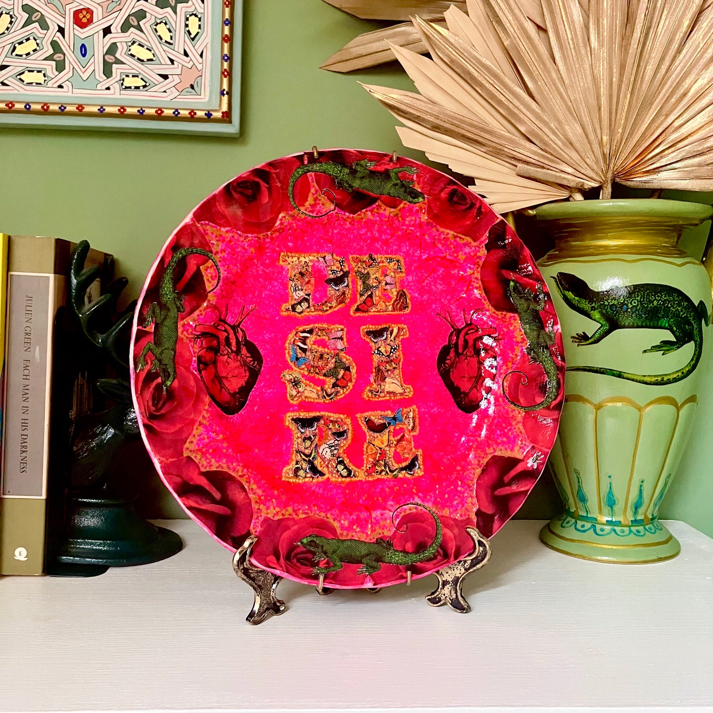 "Desire" Wall Plate by House of Frisson, featuring a collage with the word "desire" written with moths, and a frame of red roses and green lizards, on a hot pink background. Plate on a plate stand, resting on a shelf.