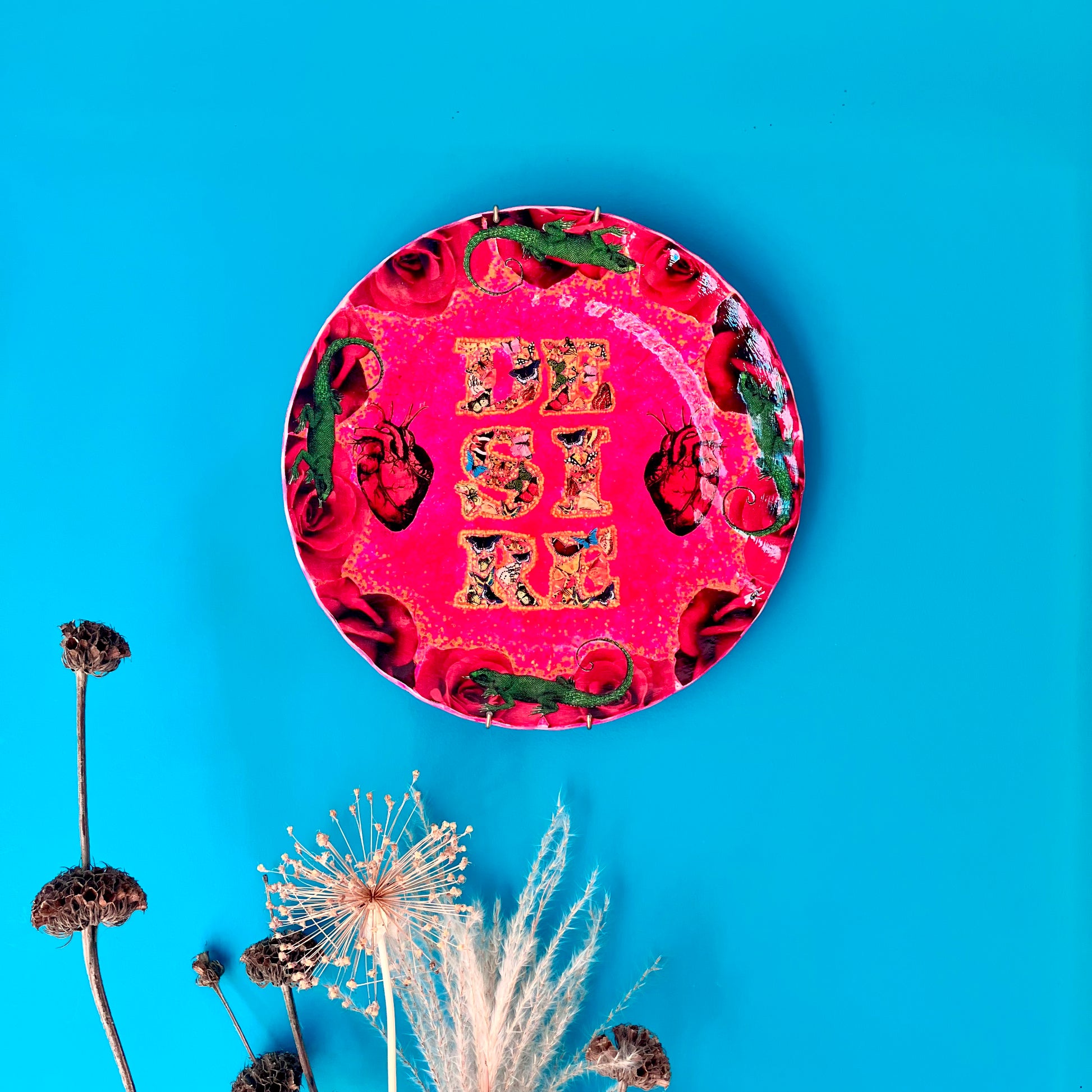 "Desire" Wall Plate by House of Frisson, featuring a collage with the word "desire" written with moths, and a frame of red roses and green lizards, on a hot pink background. Plate hanging on a blue wall.