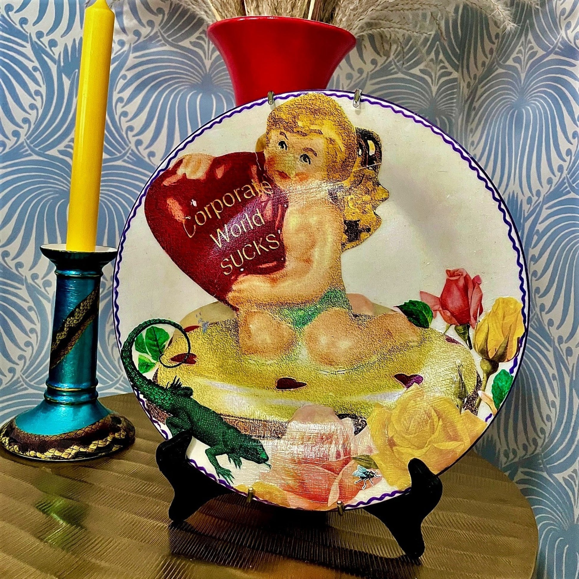 House of Frisson "Corporate World Sucks" Off White Wall Plate Front. Collage artwork featuring a kitsch angel figure holding a heart, among roses, a lizard, and a fly. Plate on a plate stand, resting on a table.