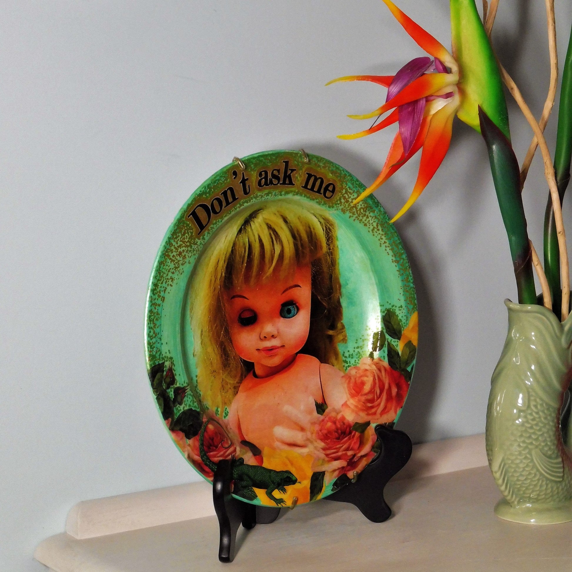 House of Frisson "Don't Ask Me" Green Wall Plate Front. Collage artwork featuring a vintage doll with funny face, surrounded by roses, and a lizard. Showing plate on a plate stand, resting on a side table.