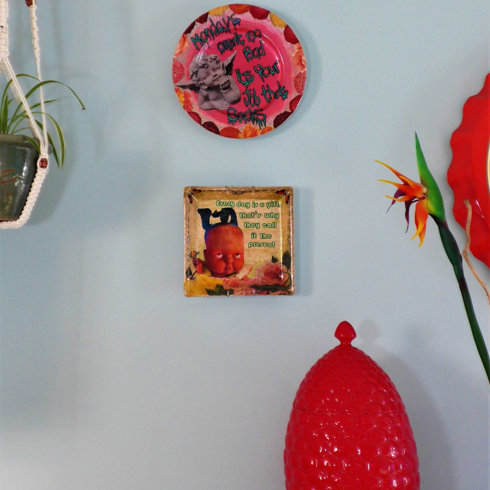 House of Frisson "Every Day Is A Gift, That's Why They Call It The Present" Beige Wall Plate. Collage artwork on upcycled plate, featuring a doll rolling  eyes, flowers, and a lizard. Showing plate hanging on wall with other plates.