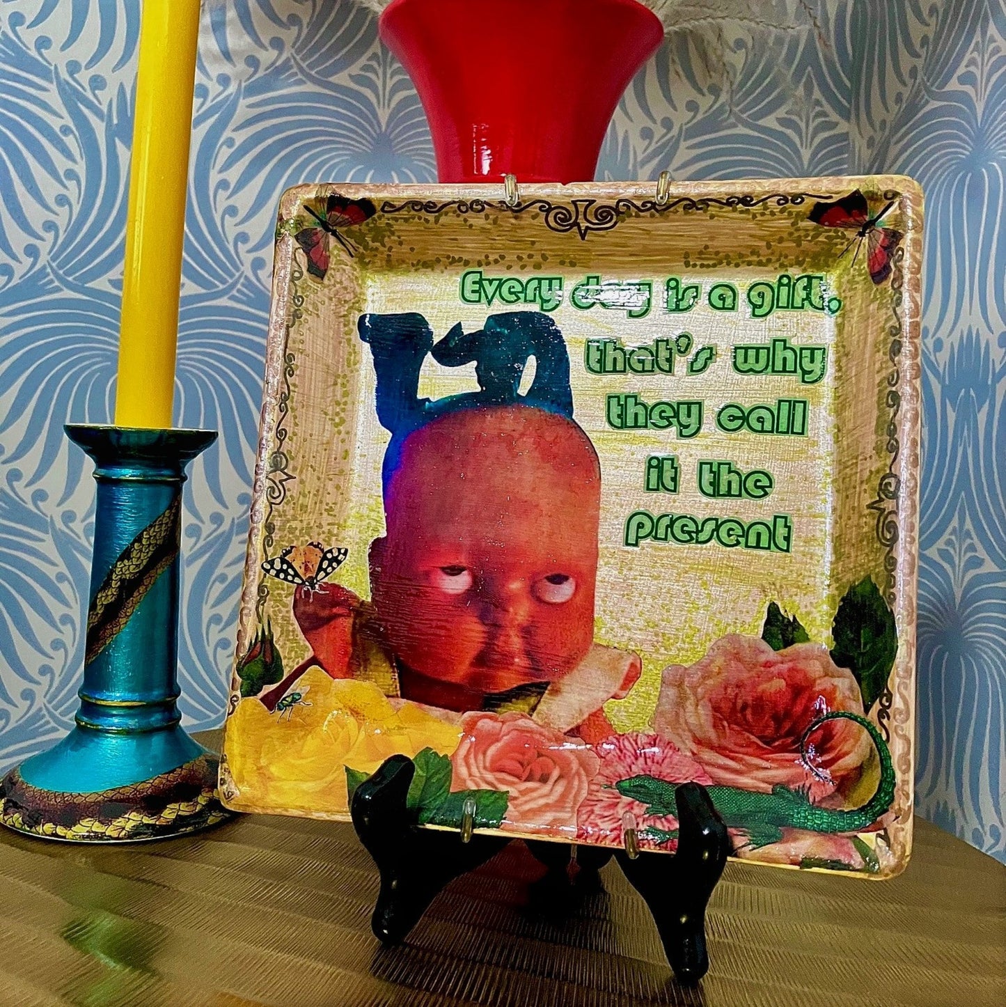 House of Frisson "Every Day Is A Gift, That's Why They Call It The Present" Beige Wall Plate. Collage artwork on upcycled plate, featuring a doll rolling  eyes, flowers, and a lizard. Plate on a plat stand, resting on a table.