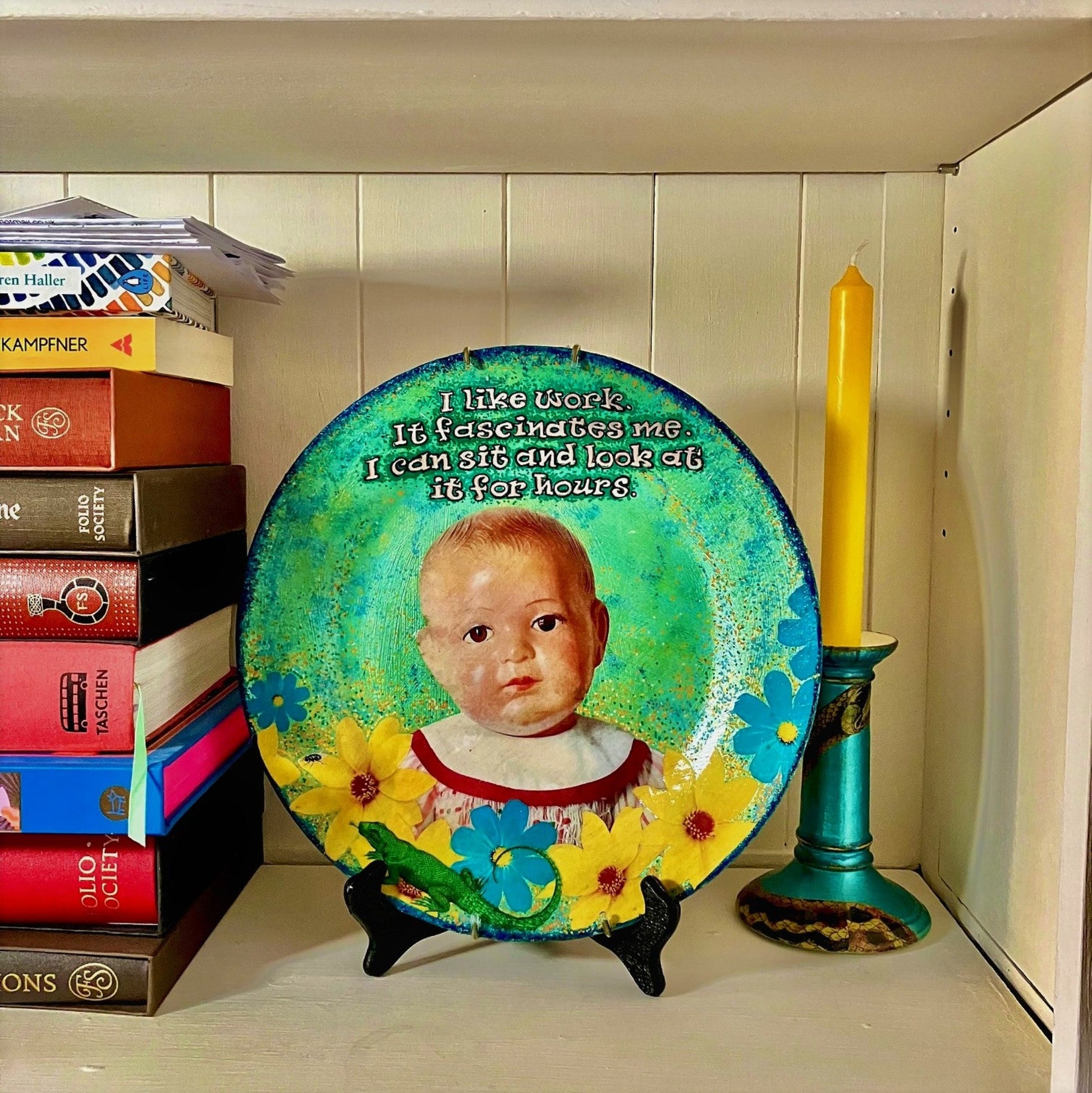 House of Frisson "I Like Work It Fascinates Me I Can Sit And Look At It For Hours" Green Wall Plate front. Collage artwork featuring a vintage male doll looking miserable among flowers, a lizard, and a fly, on a green background. Plate on a plate stand, resting on a bookshelf.
