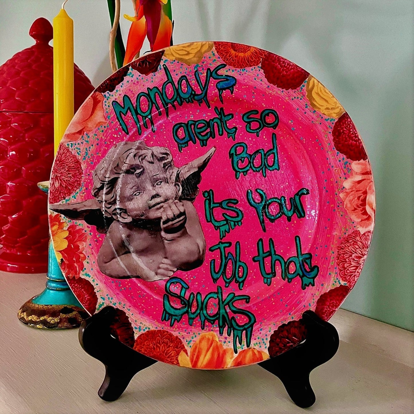 House of Frisson "Mondays Aren't So Bad, It's Your Job That Sucks" Pink Wall Plate front. Collage artwork featuring an angel statue framed with flowers, on pink background. Plate on a plate stand, resting on a side table.