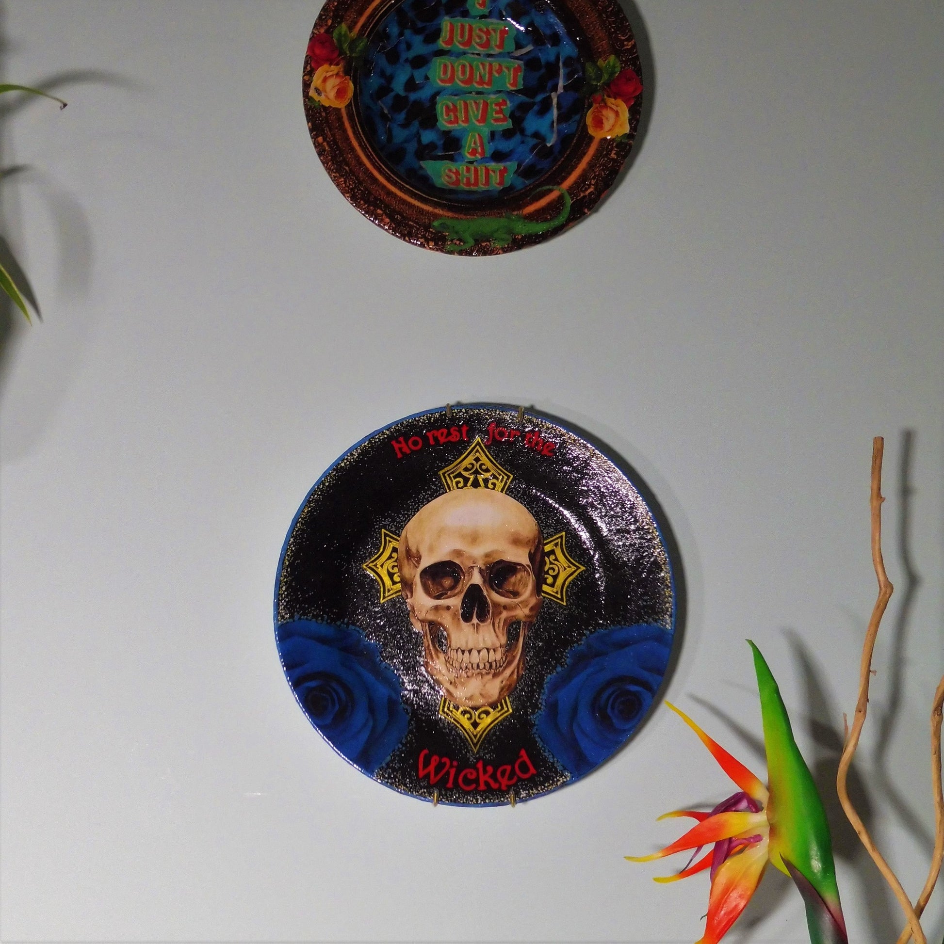 House of Frisson "No Rest For The Wicked" Black Wall Plate front. Collage artwork featuring a skull, a cross, and blue roses, on a black background. Plate hanging on a wall among other plates.