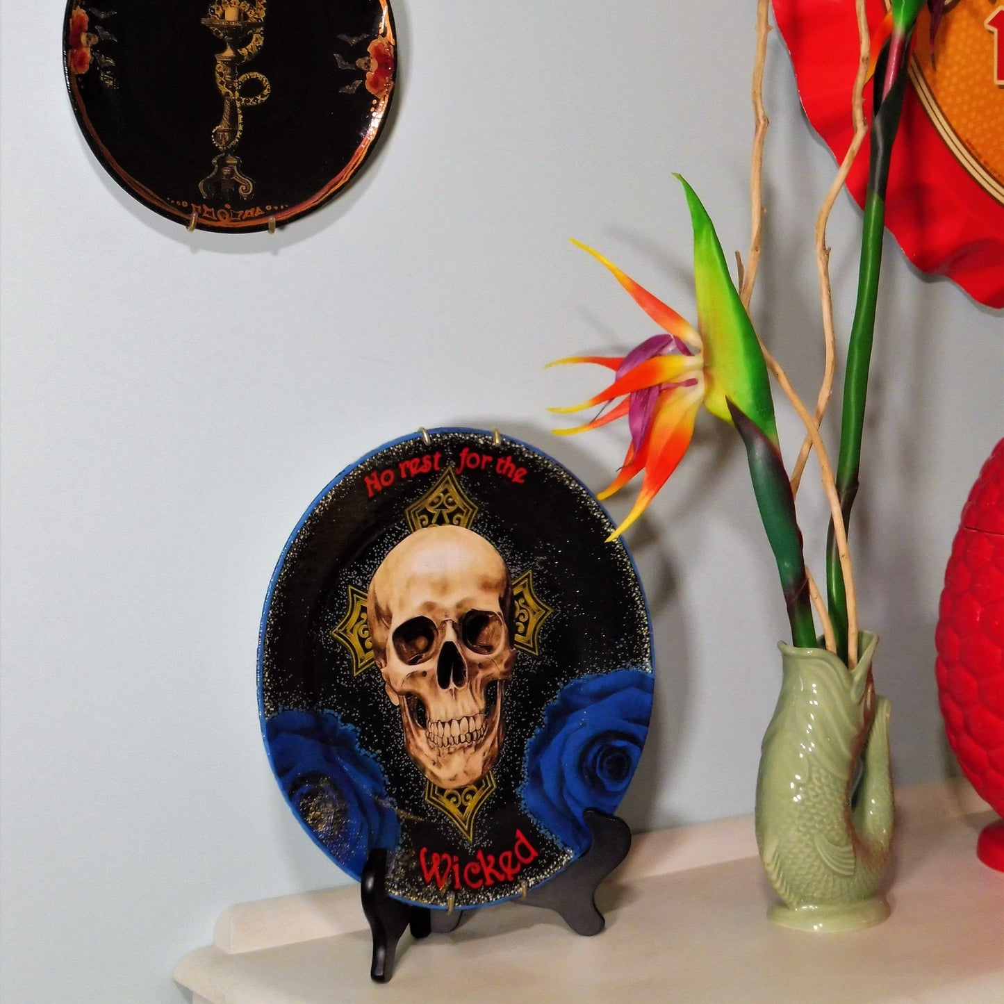 House of Frisson "No Rest For The Wicked" Black Wall Plate front. Collage artwork featuring a skull, a cross, and blue roses, on a black background. Plate on plate stand, resting on a side table.