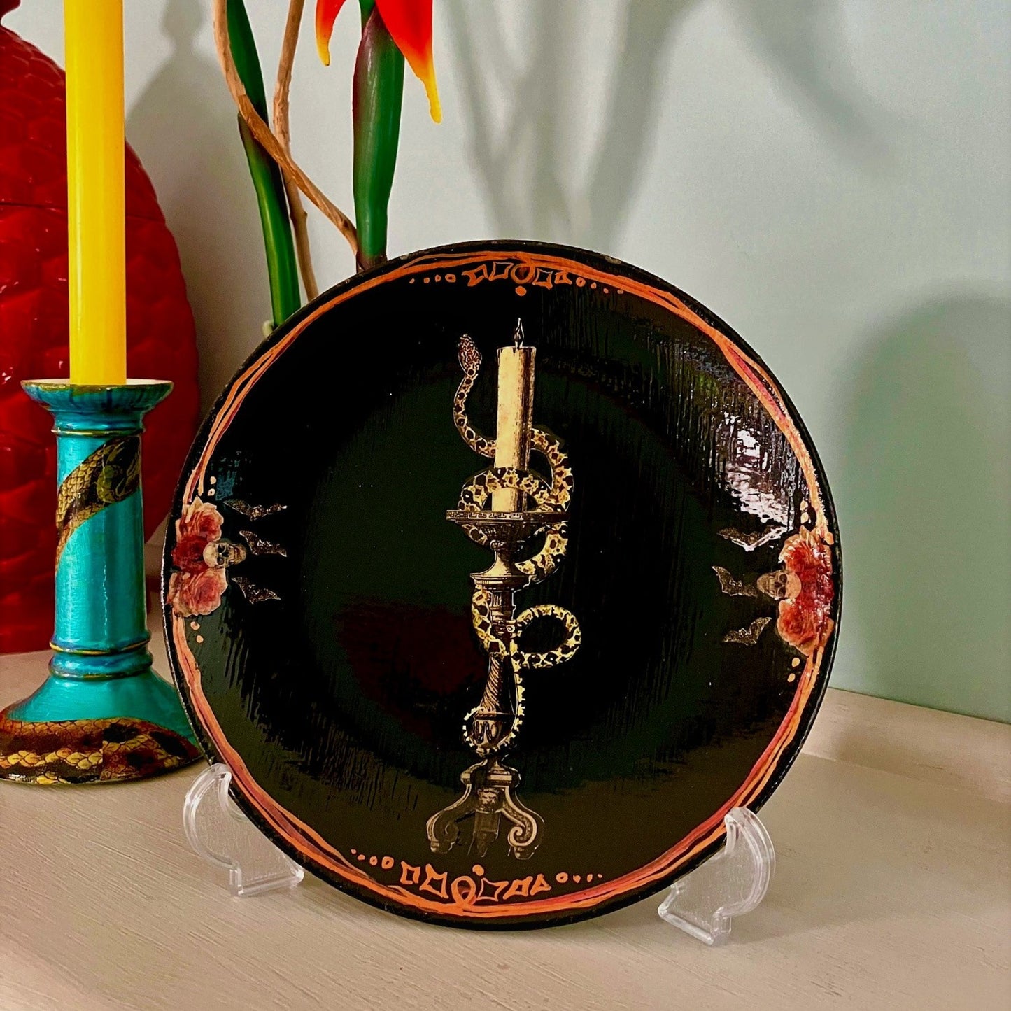 House of Frisson "Snake On Candlestick" Black Wall Plate front. Collage artwork featuring a snake wrapped around a candlestick, with skulls and bats details, on a black background. Plate on a plate stand, resting on a table.
